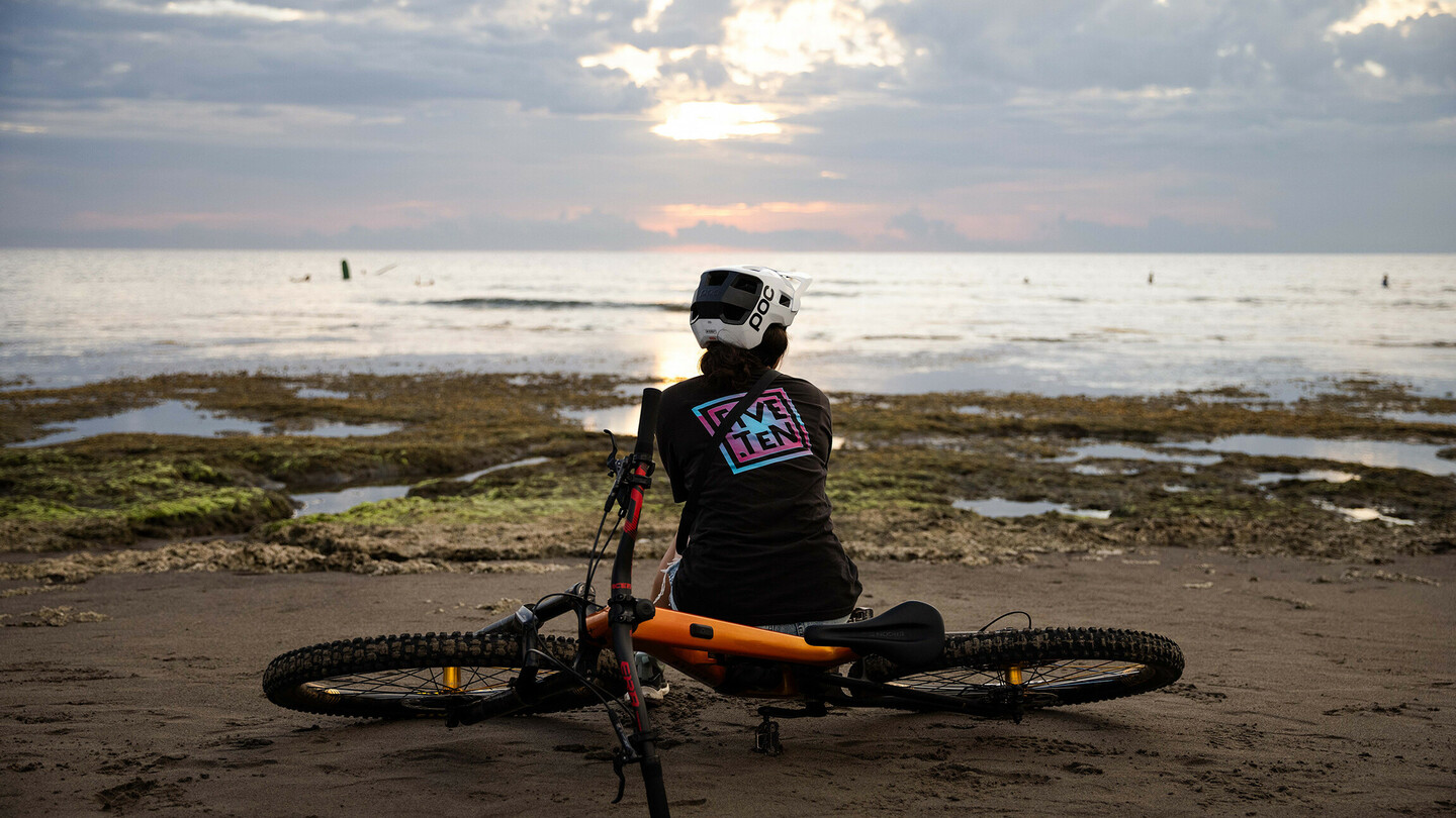 A person wearing a helmet sits on the beach next to a mountain bike, looking out at the sea as the sun sets behind the clouds. The atmosphere is calm and relaxed.