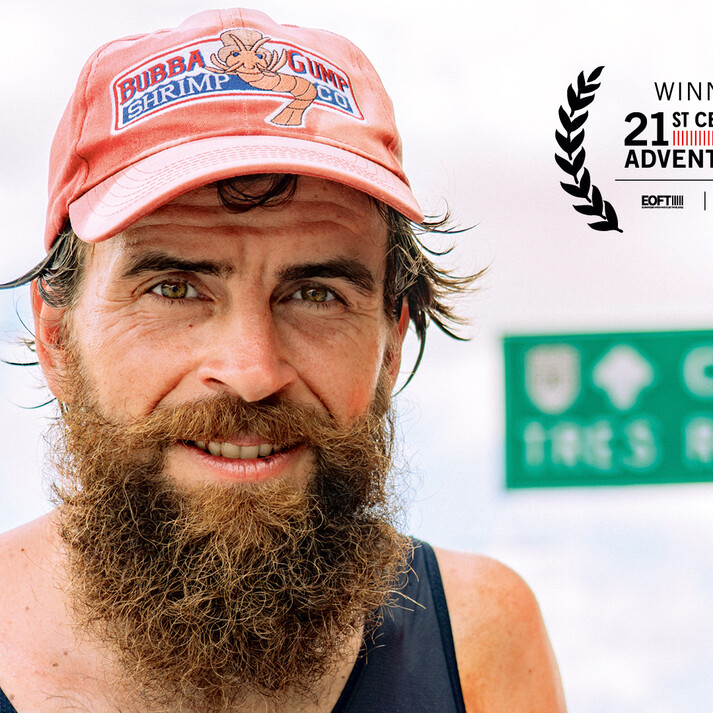 Portrait of Jonas Deichmann, a man with a thick beard and red cap featuring the Bubba Gump Shrimp Co. logo. He wears a dark tank top.